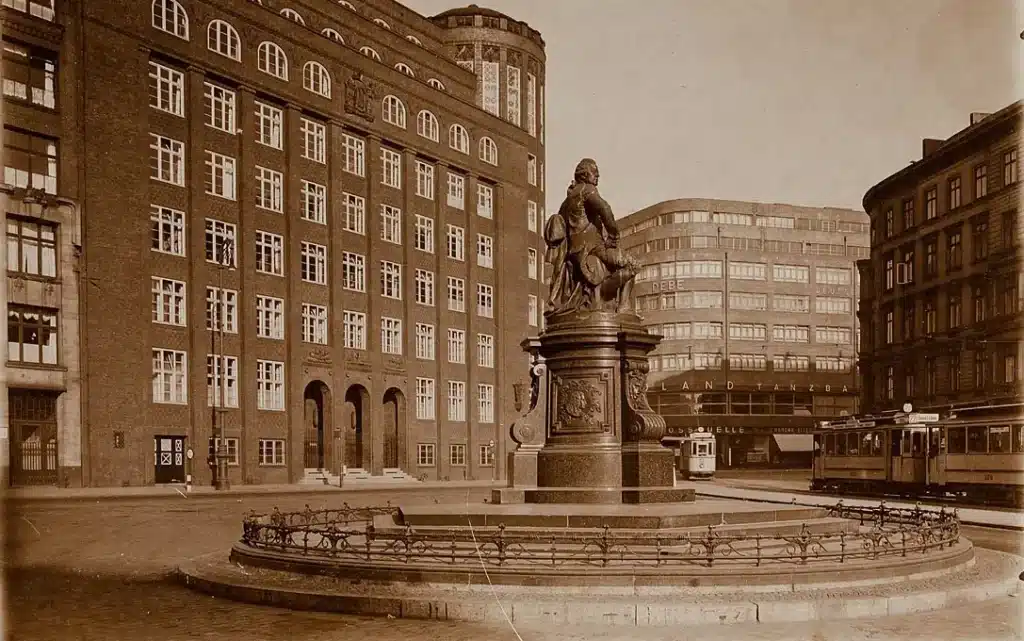 Dieses historische Foto zeigt den Hamburger Gänsemarkt im Jahr 1932 – ein faszinierender Blick zurück in eine fast vergessene Zeit. Die Aufnahme ist in sepia getönt und vermittelt das Gefühl von Geschichte, Ruhe und städtischer Ordnung.
Im Zentrum des Bildes steht das eindrucksvolle Lessing-Denkmal, auf einem massiven Sockel mit mehreren kunstvoll verzierten Stufen und Umrandungen. Die Statue zeigt Gotthold Ephraim Lessing, sitzend und nachdenklich, mit einem Buch in der Hand – ein Symbol der Aufklärung und des freien Denkens. Umgeben ist das Denkmal von einem kunstvollen gusseisernen Geländer.