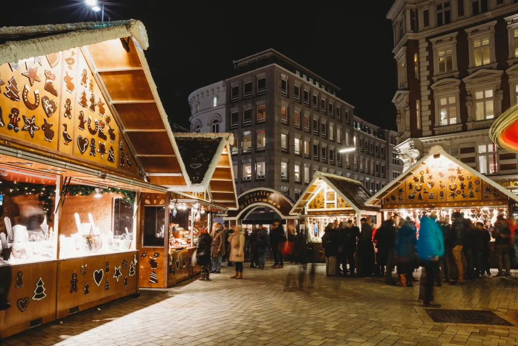 Das Bild zeigt einen stimmungsvollen Weihnachtsmarkt am Abend auf dem Hamburger Gänsemarkt. Es ist dunkel, aber der Platz ist festlich erleuchtet von den zahlreichen Lichterketten an den Weihnachtsmarktbuden. Diese kleinen Holzhäuschen sind mit Lebkuchenmotiven dekoriert – Herzen, Tannenbäume, Sterne, Hufeisen und andere winterliche Formen zieren die Wände und Dachgiebel.
In der Mitte des Bildes steht ein großes Schild mit der Aufschrift „Herzlich Willkommen“, das wie ein Torbogen zwischen zwei Buden gespannt ist. Im Hintergrund sieht man die Fassade eines modernen Bürogebäudes mit vielen Fenstern, aus denen warmes Licht scheint. Rechts im Bild ragt ein prachtvoll verziertes, historisches Gebäude mit weißen Fensterrahmen und roten Backsteinwänden empor.